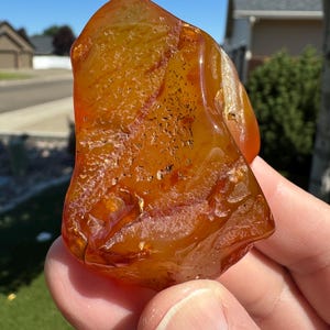 May include: A polished, amber-colored gemstone with a textured surface and internal veining. The stone is held in a hand, showcasing its translucent quality and natural variations. The gemstone's warm tones and unique patterns are highlighted in the sunlight.