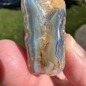 May include: A polished agate stone featuring translucent blue and grey banding with intricate, fine lines and warm, golden-brown mineral inclusions throughout the surface. The stone has a smooth, rounded texture with a glossy, reflective finish.