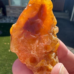 May include: A raw, translucent orange and amber-colored mineral specimen with a glossy, uneven surface texture. The stone features internal variations in color density, ranging from bright yellow-orange to deep reddish-orange hues throughout.