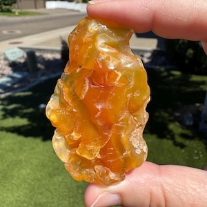 May include: A rough, amber-colored gemstone with a translucent appearance. The stone has a natural, irregular shape and is held between fingers against a sunny outdoor backdrop. The color ranges from light yellow to deep orange.