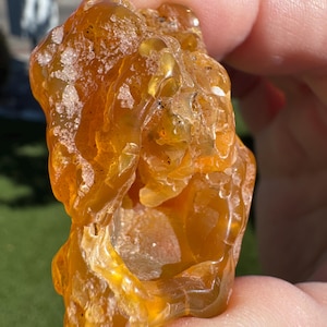 May include: A raw, amber-colored gemstone with a rough, textured surface. The stone has a translucent quality, allowing light to pass through, and is held in a person's hand. The gemstone's color ranges from light yellow to deep orange.