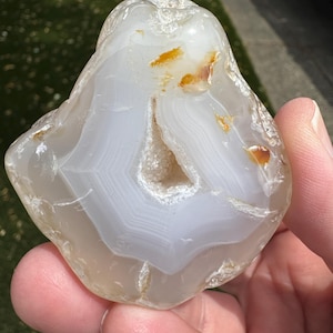May include: A polished agate stone featuring translucent white and pale gray concentric banding. The center contains a small, hollow geode cavity lined with tiny, sparkling white crystals. Several small, vibrant orange inclusions are visible.