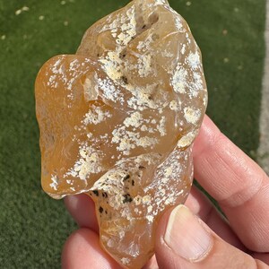 May include: A polished, irregular amber-colored agate stone featuring translucent sections, a smooth waxy texture, and scattered patches of white, crusty mineral inclusions along with small dark black specks across its surface.