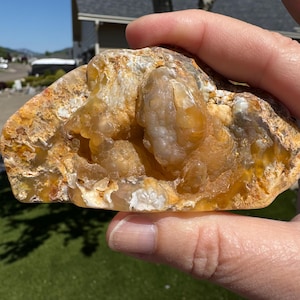 May include: A hand holds a geode with a rough, textured exterior. The geode's outer layer is a mix of tan, orange, and white hues. The interior reveals a cluster of crystalline formations in shades of yellow and white, creating a natural, earthy aesthetic.