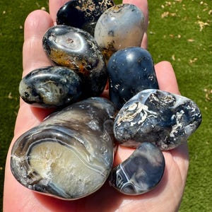 May include: A collection of polished, smooth stones in shades of black, gray, and translucent white. The stones feature varied organic patterns, including swirling bands, cloudy inclusions, and dark, glossy surfaces with reflective highlights.