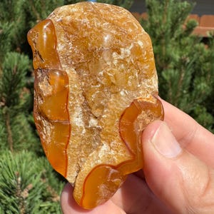 May include: A polished, irregularly shaped stone with a warm, amber color. The stone features bands of rich, orange-red hues that create a striking contrast against the lighter, translucent areas. The surface has a smooth, glossy finish.