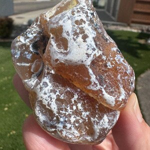 May include: A raw, translucent amber-colored stone featuring irregular white, opaque mineral inclusions across its surface. The rock has a smooth, polished texture with visible internal depth and varied brown and golden hues throughout its structure.