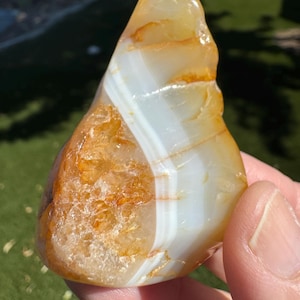 May include: A polished, triangular-shaped agate stone featuring translucent amber and golden-orange crystalline sections contrasted by distinct, smooth, opaque white bands that run vertically through the center of the mineral specimen.