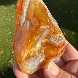May include: A polished, irregular-shaped agate stone featuring translucent layers of vibrant orange, deep amber, and creamy white. The surface displays natural banding, crystalline inclusions, and a smooth, glossy texture throughout the mineral specimen.