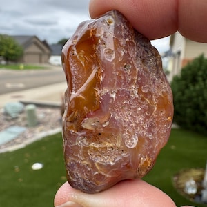 May include: A polished, natural stone with a mix of amber and reddish-brown hues. The surface has a glossy sheen and a textured appearance, with areas of translucence. The stone is held between fingers, showcasing its organic shape and color variations.