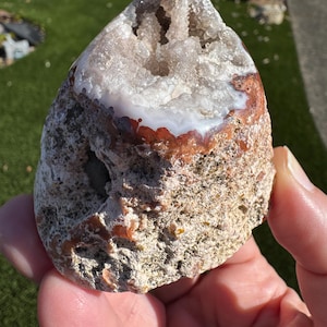 May include: A geode with a rough, textured exterior in shades of beige, brown, and orange. The interior reveals a sparkling crystal formation in white and light brown. The geode is held in a hand.