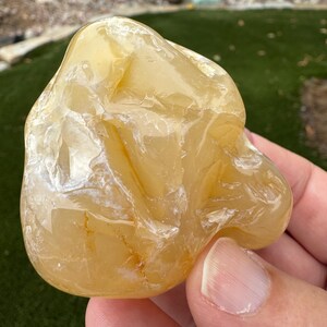 May include: A polished, translucent yellow stone with an irregular, organic shape. The surface exhibits a smooth, glossy texture with visible internal fractures, cloudy white inclusions, and subtle golden-orange veining throughout the mineral.