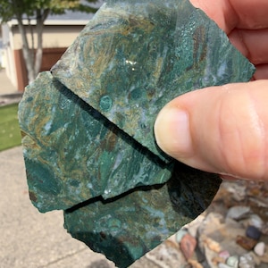 May include: Three irregular, flat pieces of polished green moss agate stone with visible brown and white inclusions. The surfaces appear wet and glossy, displaying intricate, swirling patterns of deep forest green, earthy brown, and translucent white.