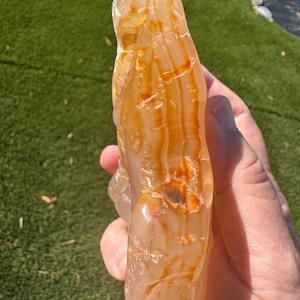 May include: A translucent, elongated stone specimen featuring a smooth, polished surface with a warm color palette of pale peach, cream, and vibrant orange bands. The mineral displays natural internal fractures and distinct, flowing color layers.