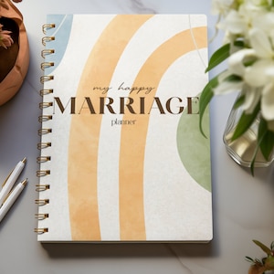 May include: A spiral-bound planner with the text "my happy MARRIAGE planner" on the cover. The cover features a watercolor design with orange, white, and green stripes. Two white pens with gold accents are next to the planner.