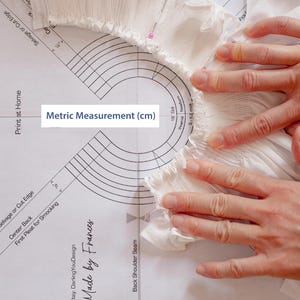 May include: A white fabric with a gathered edge is being measured with a printed guide. The guide is labelled "Metric Measurement (cm)" and has a series of concentric circles with measurements in centimetres.