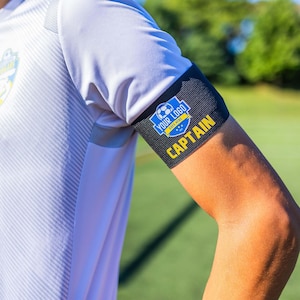 Könnte beinhalten: Schwarz-gelbes Kapitänsband mit einem Fußball-Logo und dem Text "YOUR LOGO GOES HERE" und "CAPTAIN".