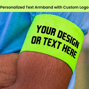 Op de afbeelding: Een neongele elastische armband met de zwarte tekst YOUR DESIGN OR TEXT HERE. De band wordt om de bovenarm gedragen over een blauwe stoffen mouw en fungeert als een opvallend accessoire dat kan worden gepersonaliseerd met logo's of tekst.