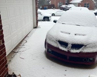 Può includere: Un'auto rossa coperta di neve parcheggiata su un vialetto. L'auto è rivolta verso la fotocamera e la parte anteriore dell'auto è coperta di neve. Il vialetto è coperto di neve e sullo sfondo c'è una casa in mattoni.