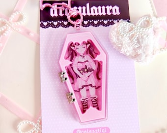 May include: A pink acrylic keychain featuring a cartoon character in a coffin-shaped design. The character has dark hair and is wearing a pink dress. The keychain has a bat-shaped clasp and the text "ursulaura" is visible on the backing card.