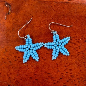 May include: A pair of light blue beaded starfish earrings with silver-colored ear wires. The earrings are made of small, round beads and are displayed on a wooden surface.