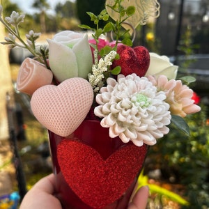 May include: A red heart-shaped tin planter filled with a bouquet of artificial flowers. The flowers include white and pink roses, a white chrysanthemum, and a green rose. The planter has a red glitter heart design.