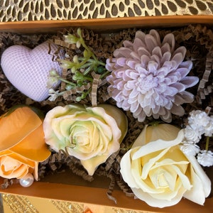 May include: A decorative gift box filled with floral-shaped soaps. The box contains a heart-shaped soap, a large chrysanthemum, and several rose-shaped soaps in shades of yellow, orange, and white. The box is lined with brown paper and has a gold exterior.