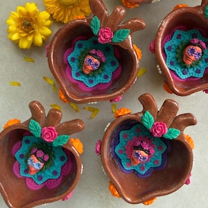 May include: Four brown ceramic bowls shaped like anatomical hearts, each featuring a central turquoise and purple floral medallion. A small orange skull with purple eye sockets sits in the center of each, accented by pink roses and green leaves.