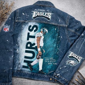 May include: A denim jacket with a painted design featuring the Philadelphia Eagles logo and the name "HURTS" in large white letters. The back of the jacket has a painted image of a football player. The NFL logo is visible.