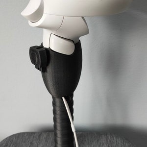 May include: A white VR controller with a black 3D printed extension handle and a white strap. The controller has a button on the side and a ring on the top.