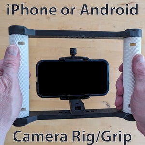 May include: A black and white camera rig/grip for a smartphone. The rig has two white handles and a black phone mount. The text "iPhone or Android" is at the top of the image and "Camera Rig/Grip" is at the bottom.