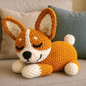 Puede incluir: Un peluche de perro corgi hecho a mano con ganchillo en naranja, blanco y negro. El perro tiene orejas grandes, una raya blanca en la cara y los ojos cerrados. El peluche está tumbado sobre una superficie gris.