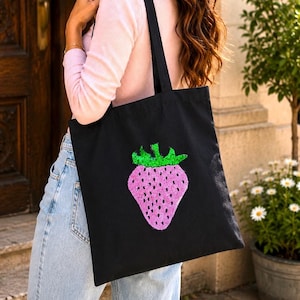 Puede incluir: Un bolso de tela negro con un gráfico grande de una fresa en el centro. La fresa está hecha de lentejuelas rosas y verdes, con pequeños puntos negros que representan las semillas, e incluye dos largas correas negras para el hombro.