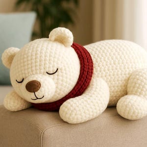 Pode incluir: Um brinquedo de pelúcia de urso de crochê creme com um cachecol vermelho. O urso tem um nariz castanho, olhos fechados e uma boca sorridente. O urso está deitado em uma superfície bege.