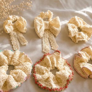 May include: A collection of six cream-colored lace hair scrunchies. Two feature long, dangling lace bows. The remaining four are circular, with two having decorative edges featuring small, colorful embroidered floral accents in pink and pastel tones.