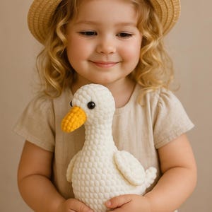 May include: A crocheted white goose plush toy with a yellow beak and feet. The toy is held by a person wearing a straw hat and a beige dress. The goose has black eyes and a textured body.