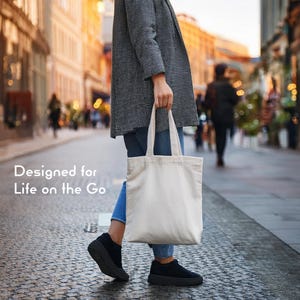 Op de afbeelding: Een effen, gebroken witte canvas tas met twee schouderbanden, aan de zijkant vastgehouden. De tekst "Designed for Life on the Go" is in het wit gedrukt aan de linkerkant van de afbeelding, tegen een wazige stedelijke straatachtergrond.