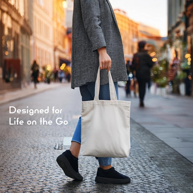Op de afbeelding: Een effen, gebroken witte canvas tas met twee schouderbanden, aan de zijkant vastgehouden. De tekst "Designed for Life on the Go" is in het wit gedrukt aan de linkerkant van de afbeelding, tegen een wazige stedelijke straatachtergrond.