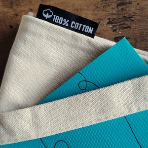 Op de afbeelding: Een beige canvas draagtas met een zwart stoffen label aan de bovenrand, voorzien van een wit katoenbol-icoon en de tekst 100% COTTON. Een helderblauwe, getextureerde kaart met zwarte lijntekeningen steekt gedeeltelijk uit de opening van de tas.
