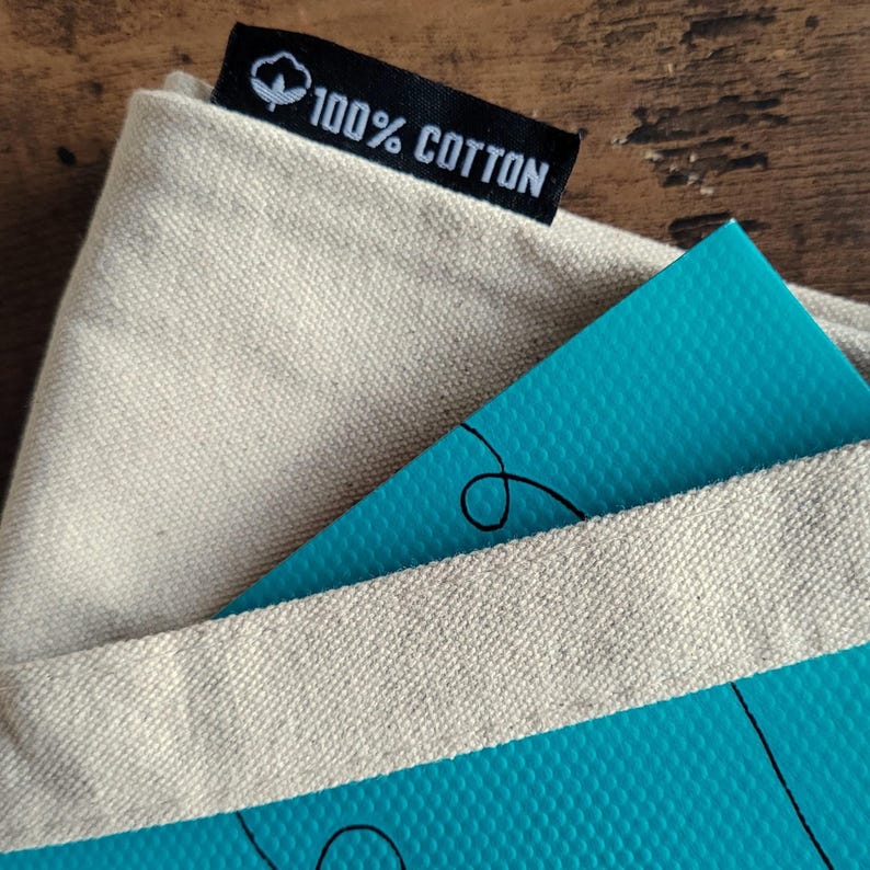 Op de afbeelding: Een beige canvas draagtas met een zwart stoffen label aan de bovenrand, voorzien van een wit katoenbol-icoon en de tekst 100% COTTON. Een helderblauwe, getextureerde kaart met zwarte lijntekeningen steekt gedeeltelijk uit de opening van de tas.