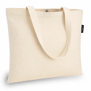 Op de afbeelding: Een rechthoekige, gebroken witte canvas tas met twee schouderbanden en een centrale metalen drukknoopsluiting. Aan de rechterzijnaad zit een klein zwart stoffen label met witte tekst, waarop de merknaam VAGABOND staat vermeld.