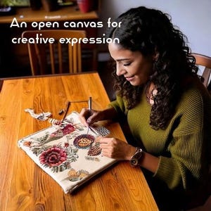 Op de afbeelding: Een vierkant stoffen tasje met een gedetailleerd geborduurd bloemenpatroon in rood-, groen- en geeltinten. De tekst "An open canvas for creative expression" is bovenaan zichtbaar. Een metalen naald wordt gebruikt om steken aan het ontwerp toe te voegen.