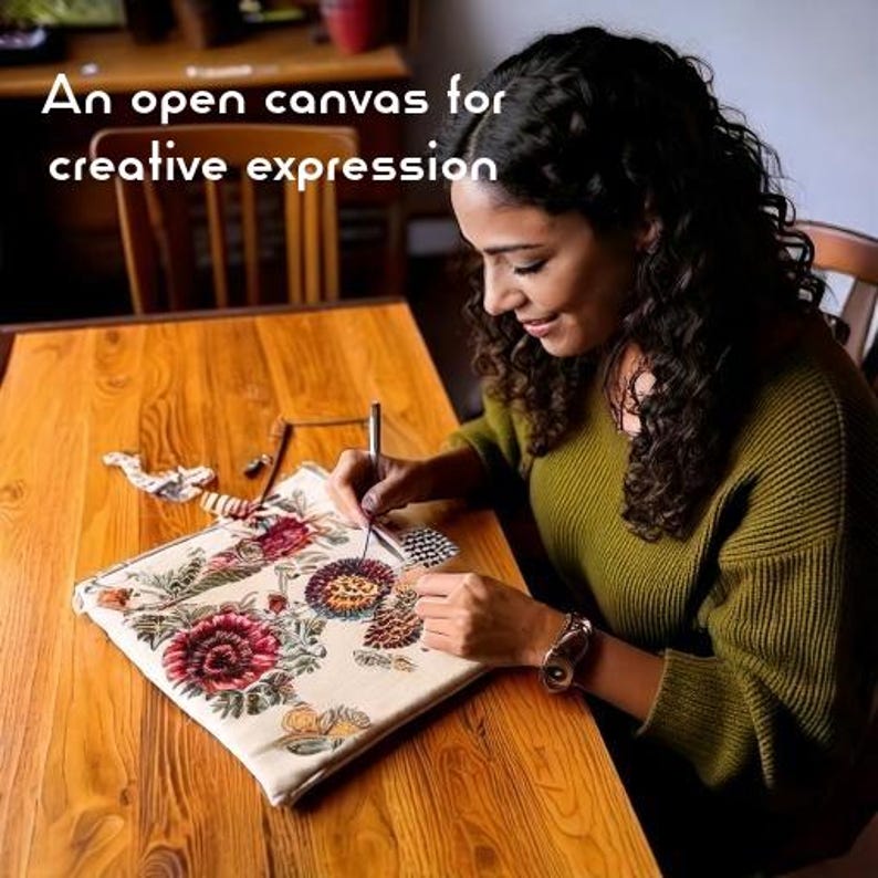 Op de afbeelding: Een vierkant stoffen tasje met een gedetailleerd geborduurd bloemenpatroon in rood-, groen- en geeltinten. De tekst "An open canvas for creative expression" is bovenaan zichtbaar. Een metalen naald wordt gebruikt om steken aan het ontwerp toe te voegen.