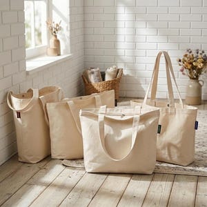 Op de afbeelding: Een collectie van vier off-white canvas draagtassen met twee schouderbanden. Elke tas heeft een rechthoekige bodem, een ruim hoofdvak en een klein rechthoekig stoffen label dat aan de zijnaad is bevestigd. De tassen zijn gemaakt van stevige stof.