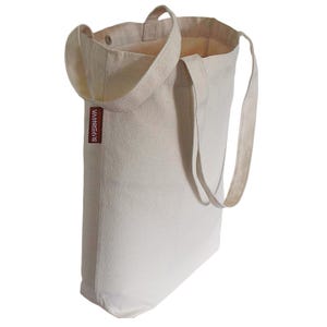 Op de afbeelding: Een gebroken witte canvas draagtas met twee lange schouderbanden en een klein rechthoekig bruin stoffen label aan de zijkant met de witte tekst SUPERNOVA. De tas heeft een open bovenkant met een zichtbare metalen drukknoop aan de binnenzijde.