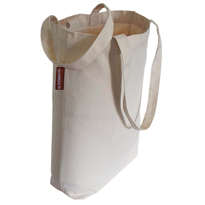 Op de afbeelding: Een gebroken witte canvas draagtas met twee lange schouderbanden en een klein rechthoekig bruin stoffen label aan de zijkant met de witte tekst SUPERNOVA. De tas heeft een open bovenkant met een zichtbare metalen drukknoop aan de binnenzijde.