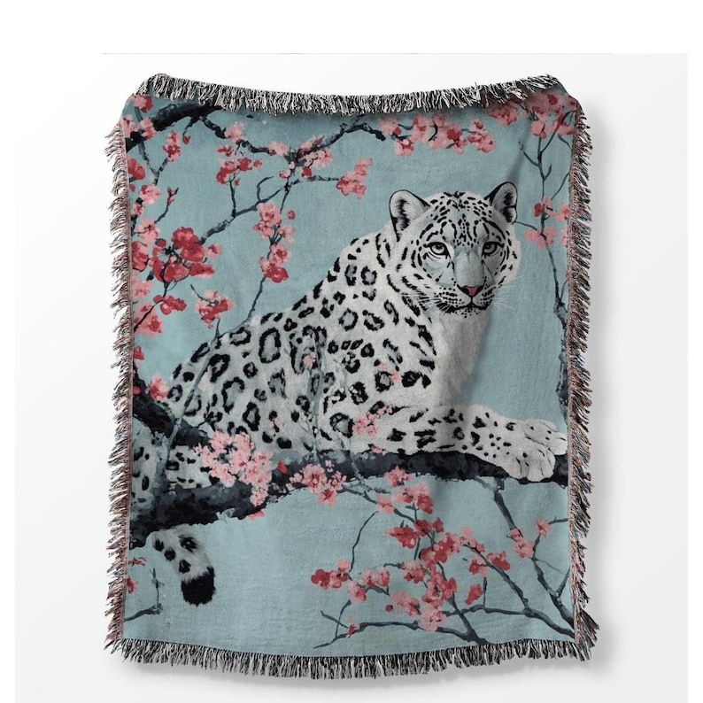 Op de afbeelding: Geweven sprei met een sneeuwluipaard dat rust op een tak met roze kersenbloesems. De sprei heeft een lichtblauwe achtergrond en een gefranjerde rand. Het sneeuwluipaard heeft witte vacht met zwarte vlekken.
