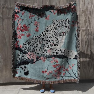 Op de afbeelding: Een geweven sprei met een sneeuwluipaard dat rust op een tak met roze kersenbloesems tegen een turquoise achtergrond. De deken heeft een gefranjerde rand en is gemaakt van een zacht, comfortabel materiaal.