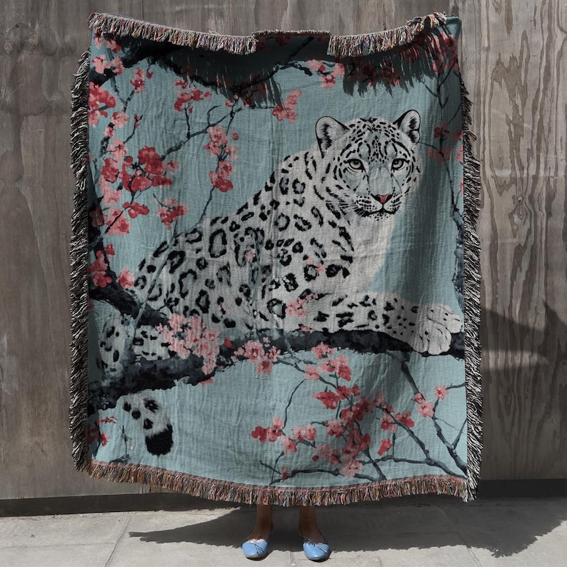 Op de afbeelding: Een geweven sprei met een sneeuwluipaard dat rust op een tak met roze kersenbloesems tegen een turquoise achtergrond. De deken heeft een gefranjerde rand en is gemaakt van een zacht, comfortabel materiaal.