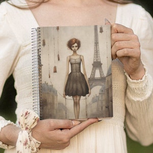 May include: A spiral bound notebook with a cover featuring a watercolor illustration of a woman in a black dress standing on a string in front of the Eiffel Tower. The background is a muted gray with hints of brown and green.